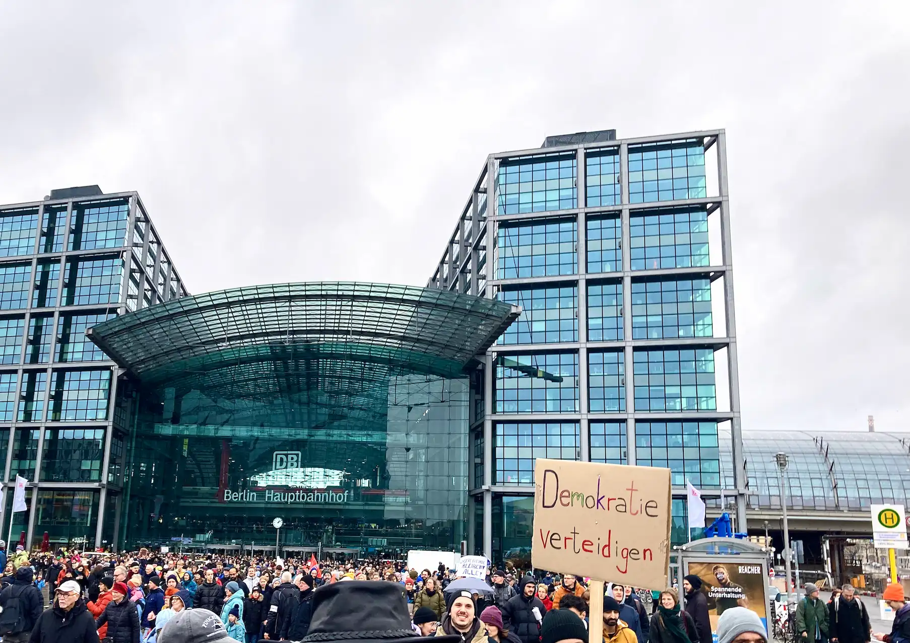 Demonstrierende Menschen vor dem Berliner Hauptbahnhof. Im Vordergrund ist ein selbst gemachtes Plakat mit der Aufschrift „Demokratie verteidigen“ zu sehen.
